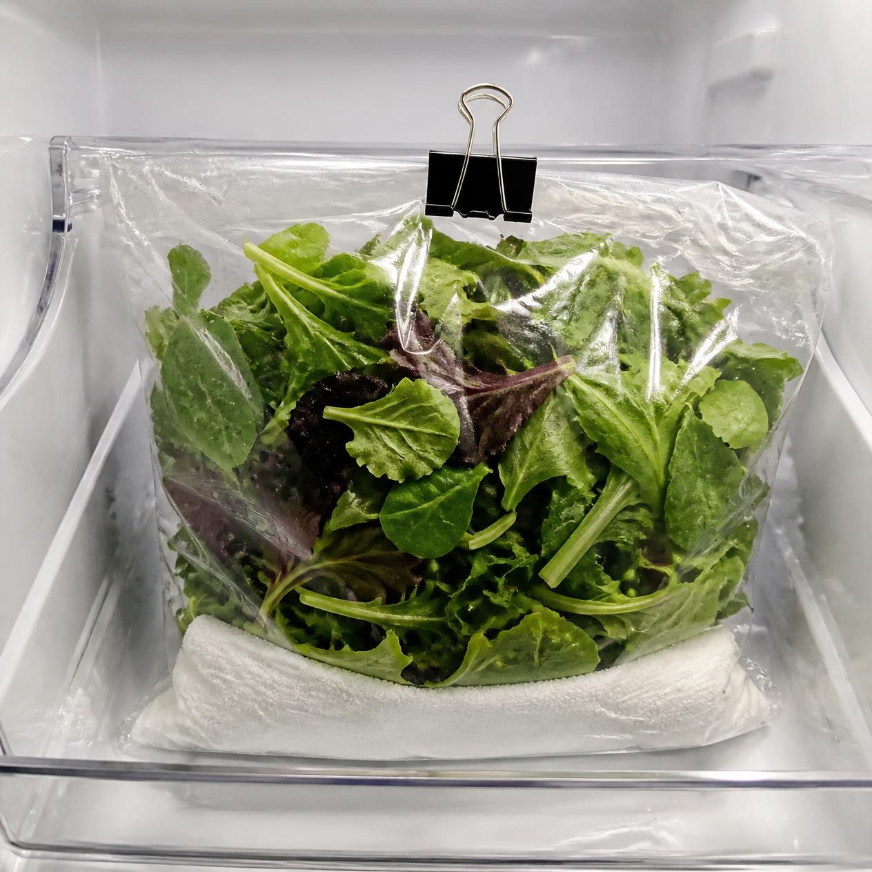A clipped bag of greens with a cloth at the bottom, in the fridge